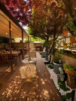 An outdoor dining area with tables and chairs, surrounded by greenery and a stone wall.