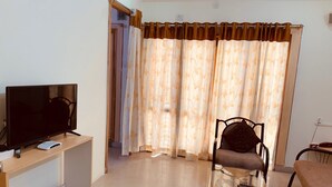 A living room with a flat-screen TV on a wooden stand, a glass coffee table, a brown armchair, and a sofa. - Khyathi Hotels (Hyderabad)