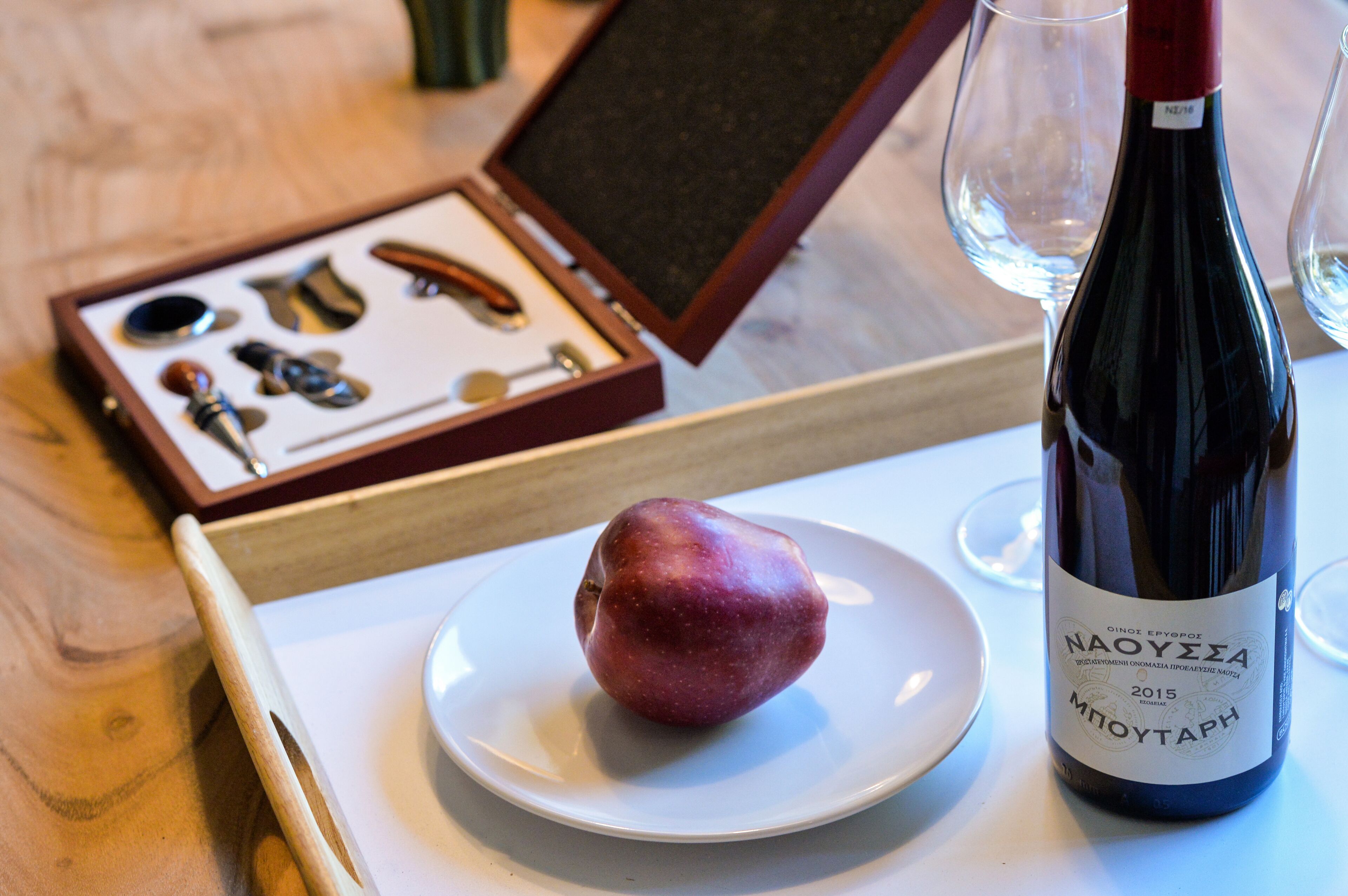A wine bottle with a label, a plate with a single fruit, and a set of wine glasses and corkscrews on a tray.