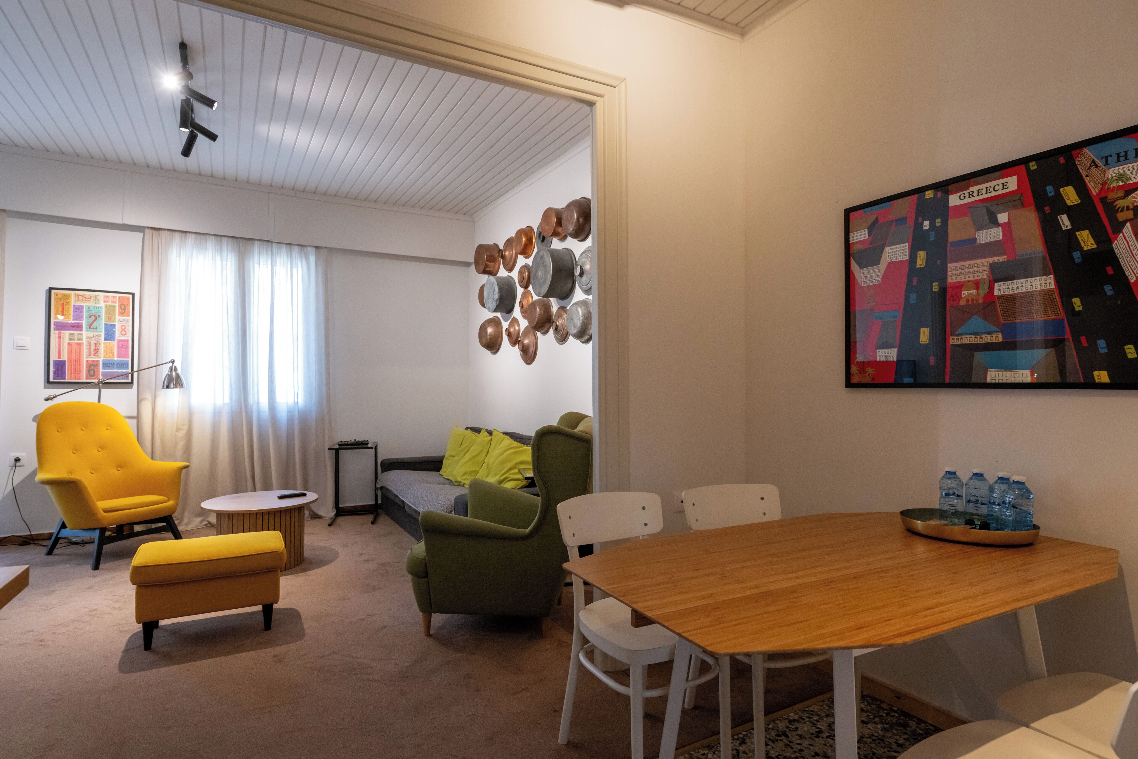 A modern living room with a wooden table, yellow and green armchairs, a sofa, and a wall art piece.