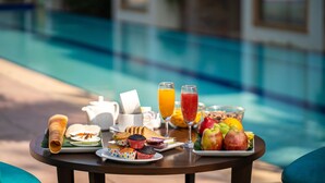 A breakfast table with a variety of foods and drinks, including a rolled pastry, muffins, and a fruit bowl, by a poolside. - Ramya Resort & Spa (Udaipur)
