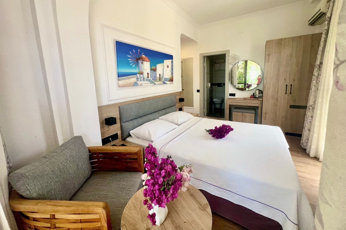 A bedroom with a bed, a wooden side table with a vase of flowers, a painting of a coastal scene, a wooden wardrobe, and a bathroom visible through an open door.
