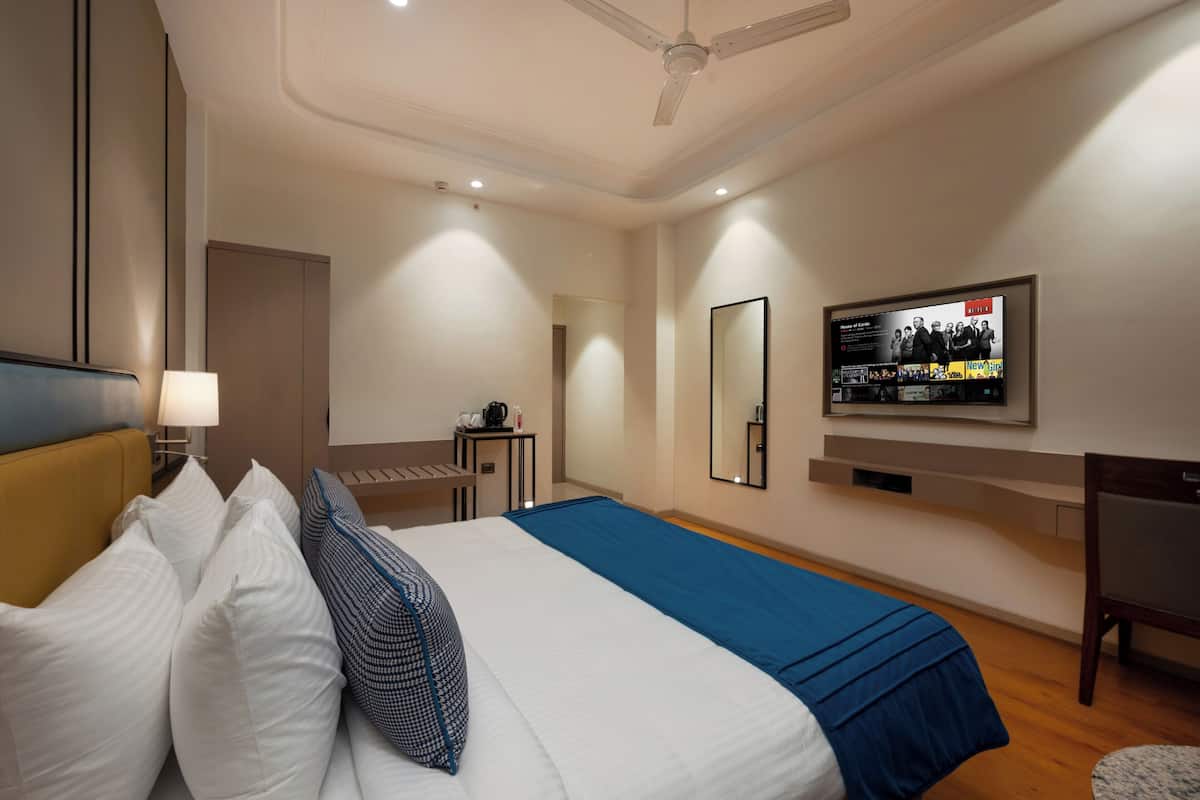 A hotel room with a bed, a desk, a chair, a TV mounted on the wall, and a ceiling fan.
