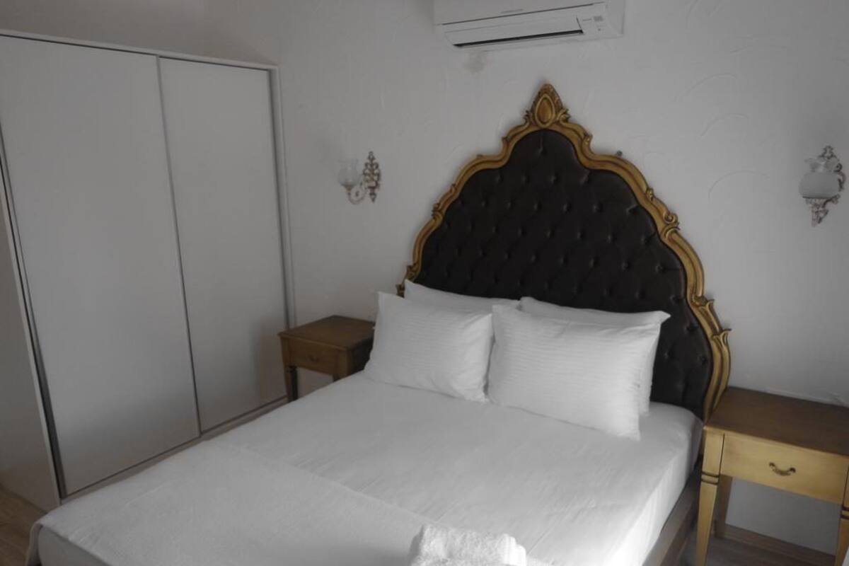 A neatly made bed with white linens, a dark upholstered headboard, and a wooden nightstand with a drawer.