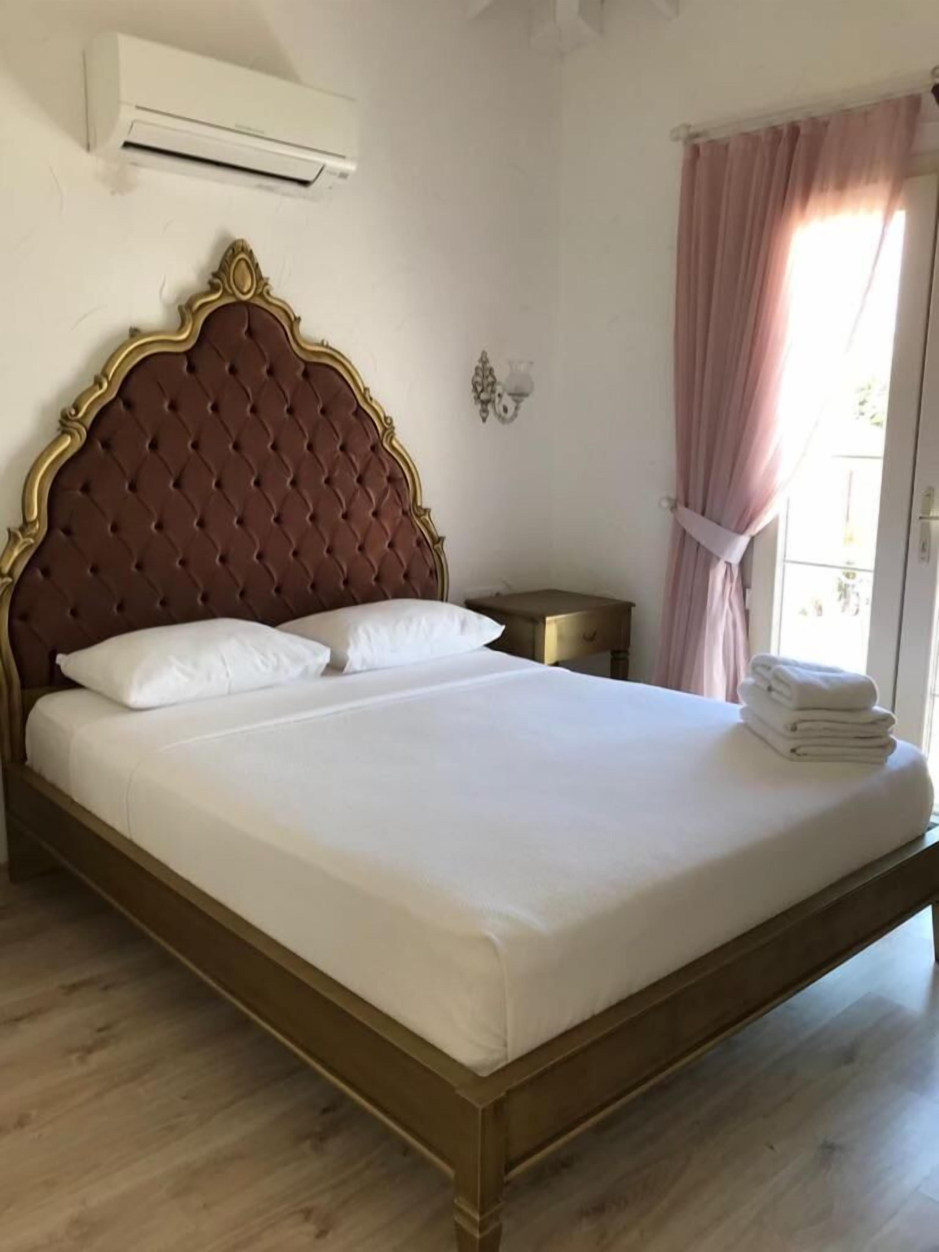 A neatly made bed with a wooden frame and a tufted headboard, a bedside table, and a stack of folded towels.