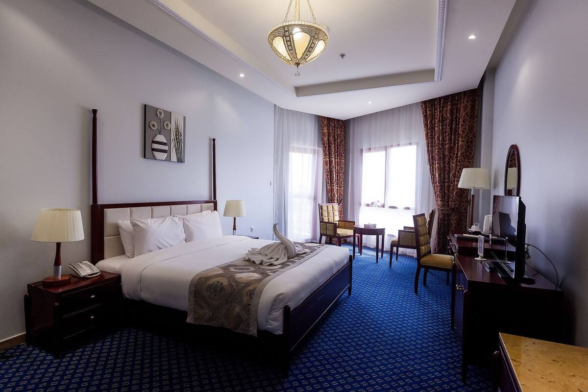 A hotel room with a large bed, bedside tables, a desk with a chair, a television, and a chandelier.