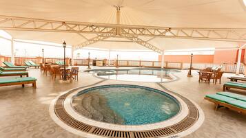 A covered swimming pool area with lounge chairs and tables, a central pool, and a tiled floor.