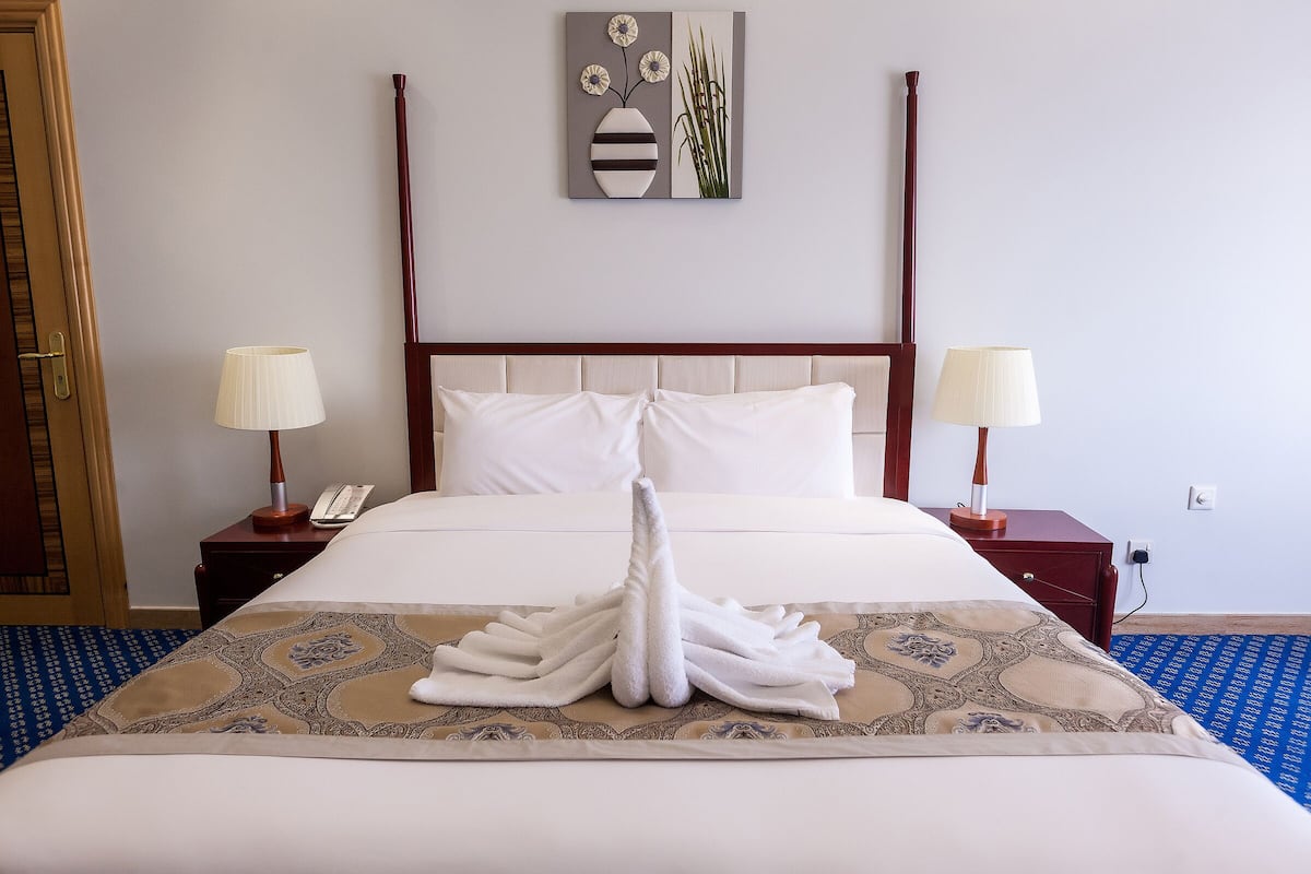 A neatly made bed with a towel arrangement, a bedside table with a lamp, a phone, and a wall art piece above the bed.