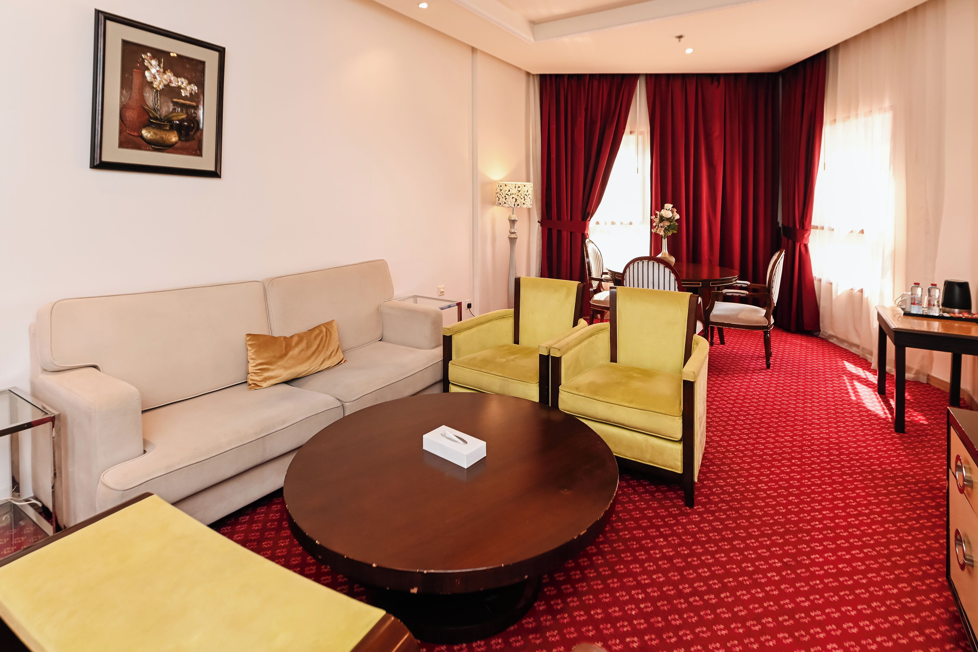 Une chambre d’hôtel avec un canapé, des fauteuils, une table basse, une table à manger avec des chaises et une moquette rouge.