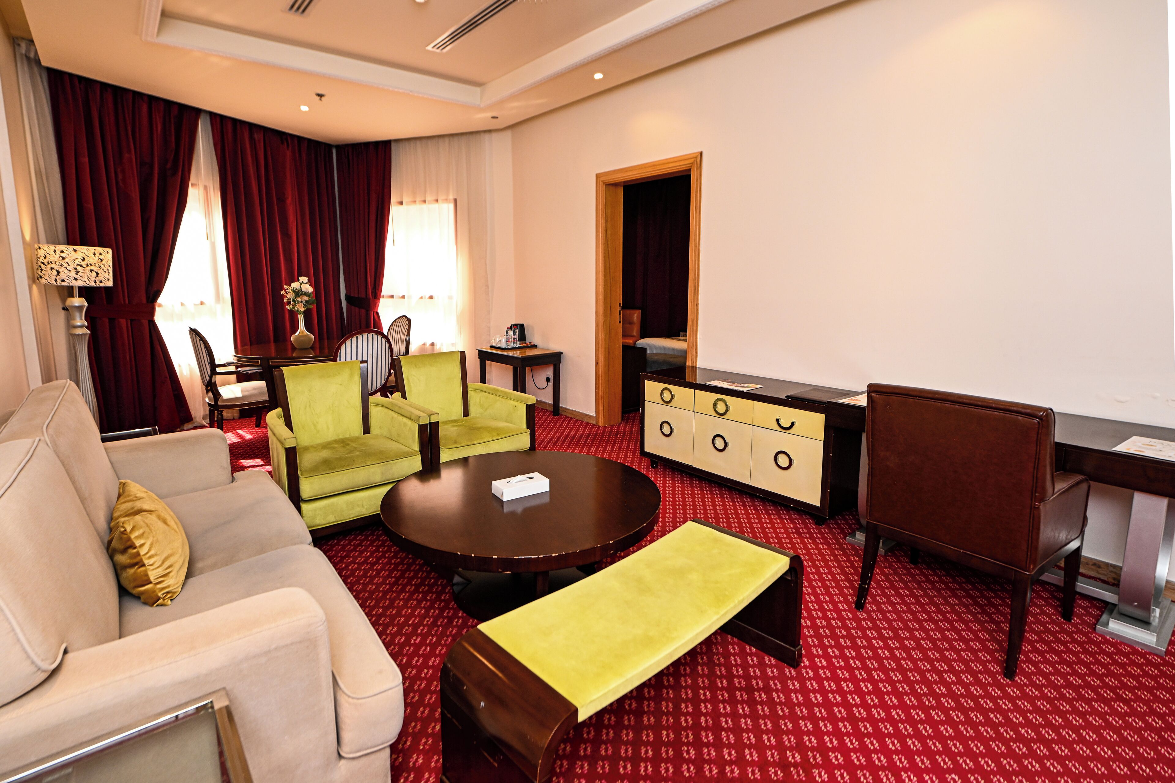 A hotel room with a red carpet, a beige sofa, a green armchair, a wooden coffee table, a brown leather chair, and a wooden cabinet.