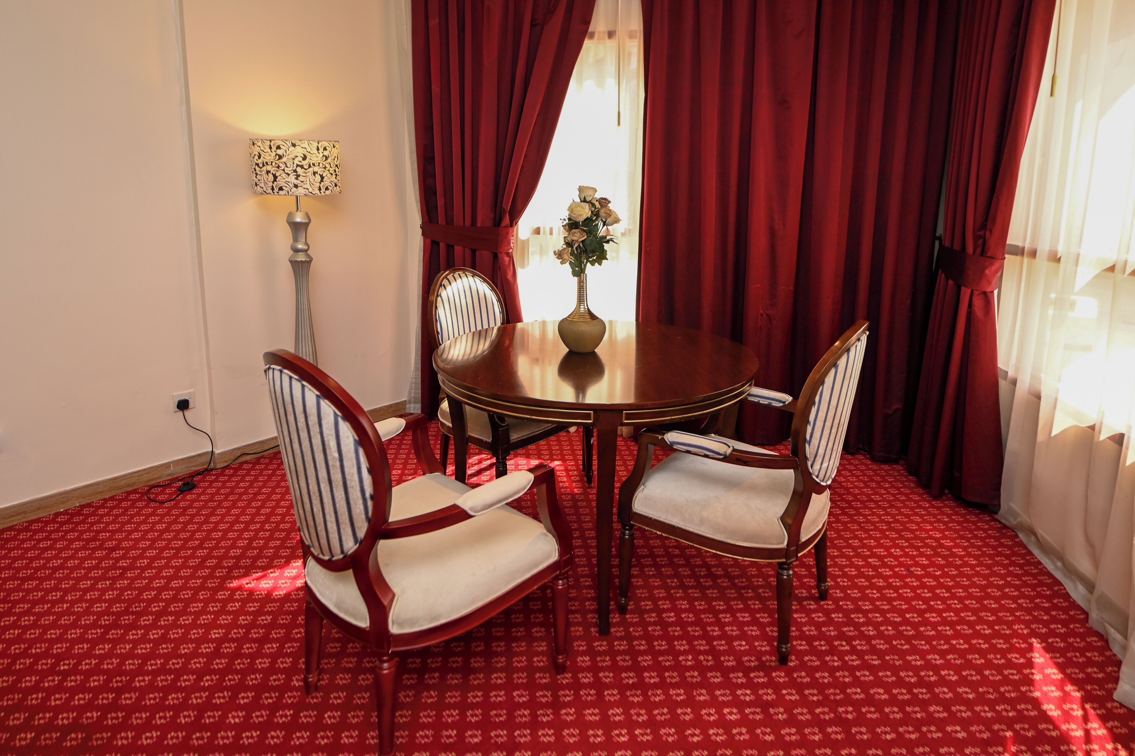 A dining area with a round table, four chairs, a vase with flowers, red curtains, and a lamp.