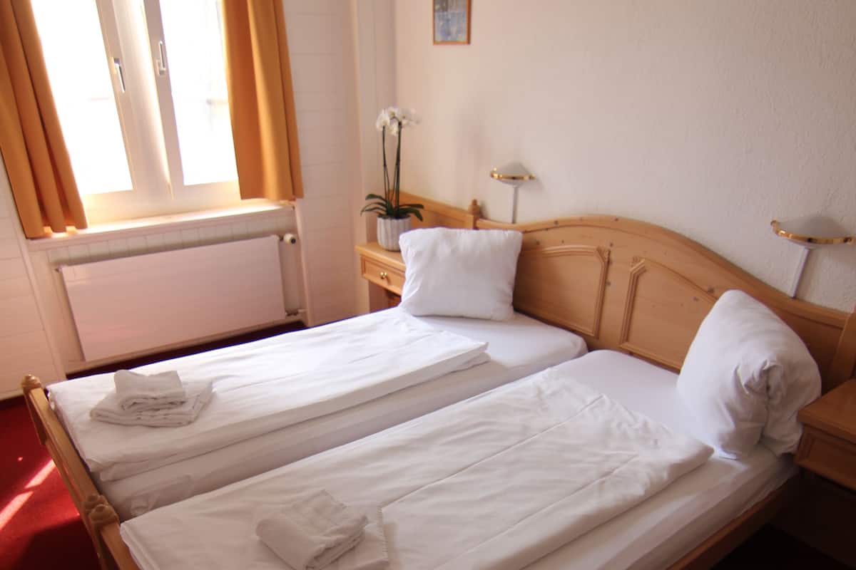 Een tweepersoonsbed met witte lakens en handdoeken, een houten hoofdbord, een nachtkastje met een lamp en een raam met gordijnen.