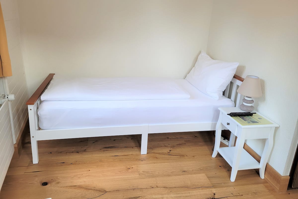 Een wit bed met een houten hoofdbord, een witte nachtkastje met een lamp, en een houten vloer.