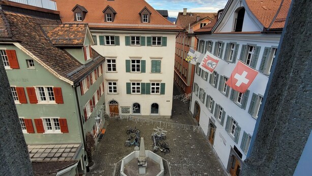 A narrow street with buildings on either side, a central fountain, and flags. - Hotel Zunfthaus zur Rebleuten (Chur)