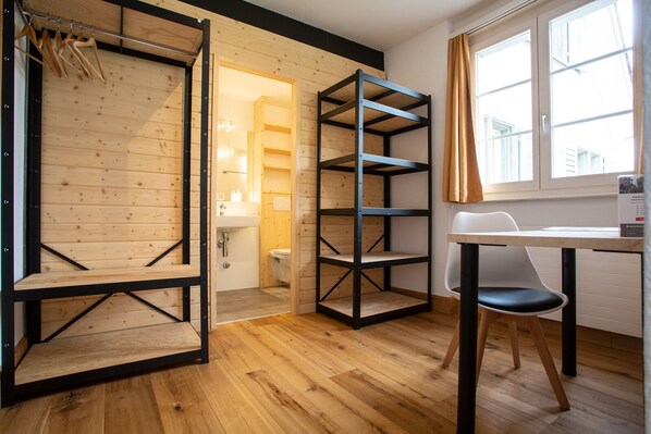 A room with a wooden floor, a white desk with a chair, a black metal shelving unit, and a wooden door leading to a bathroom.