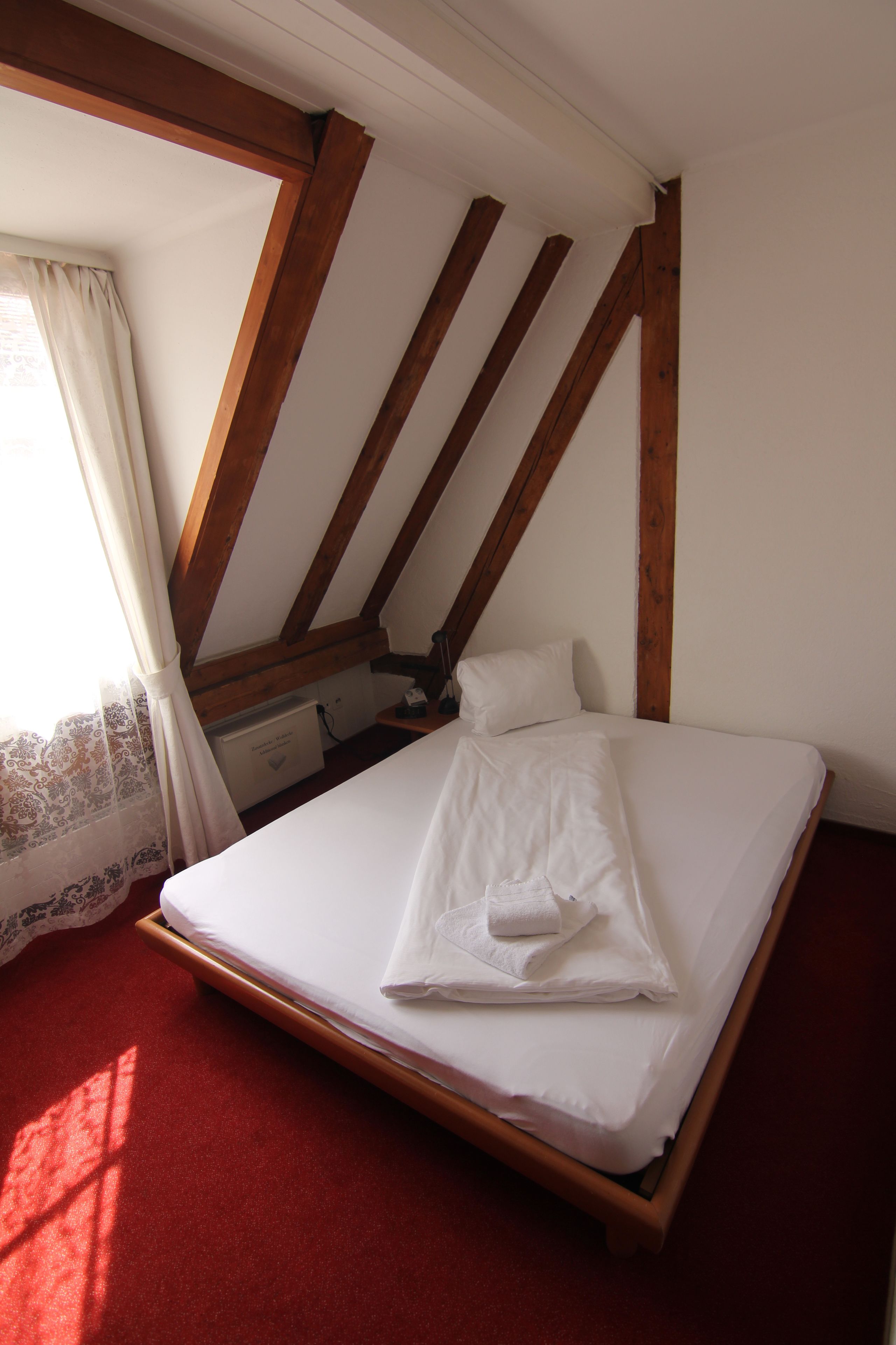 Ein Einzelbett in einem Zimmer mit Holzbalken, einem roten Teppich und einem Fenster mit Vorhang.