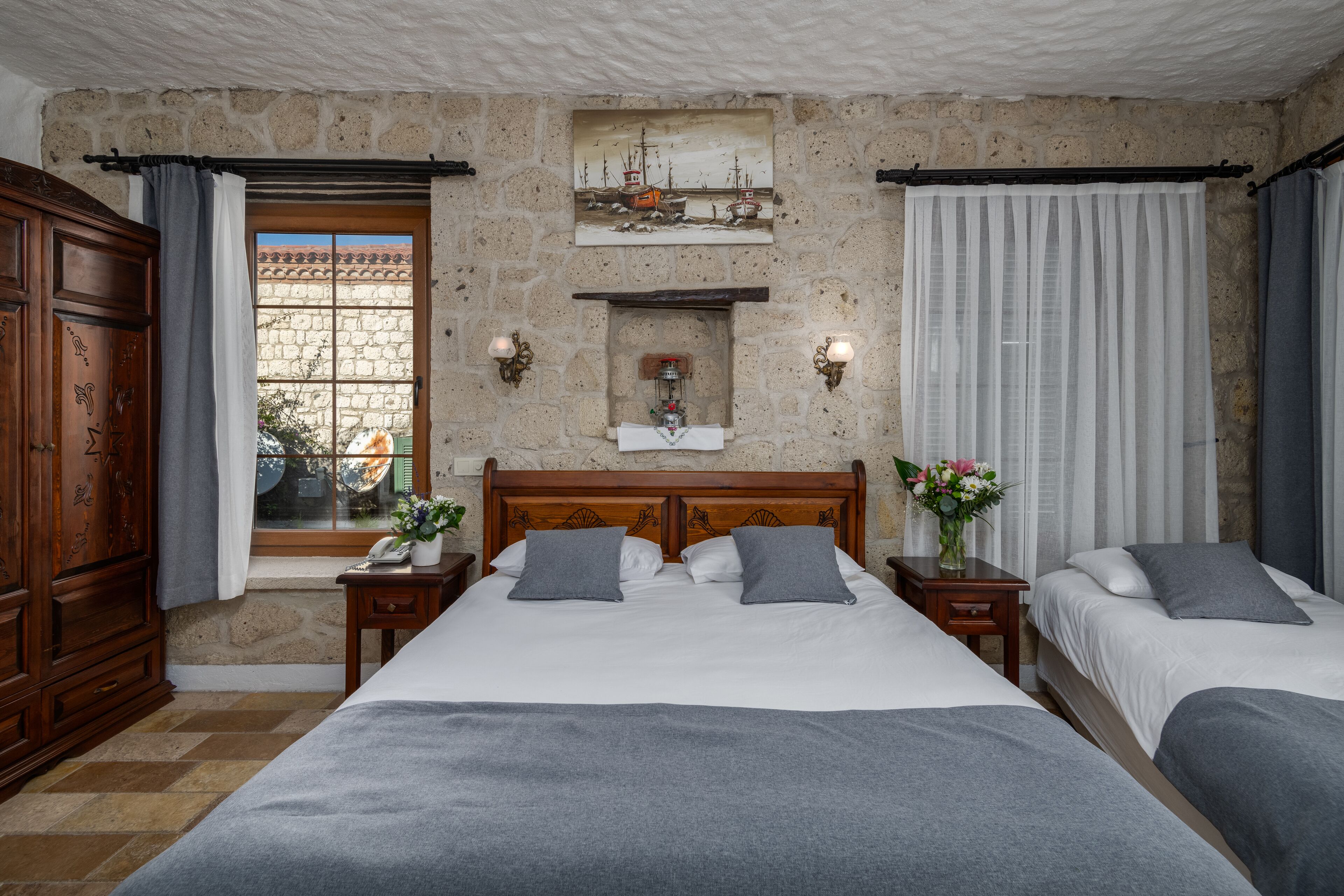 A hotel room with a stone wall, a bed with grey bedding, a wooden headboard, a nightstand with a lamp, and a window with curtains.