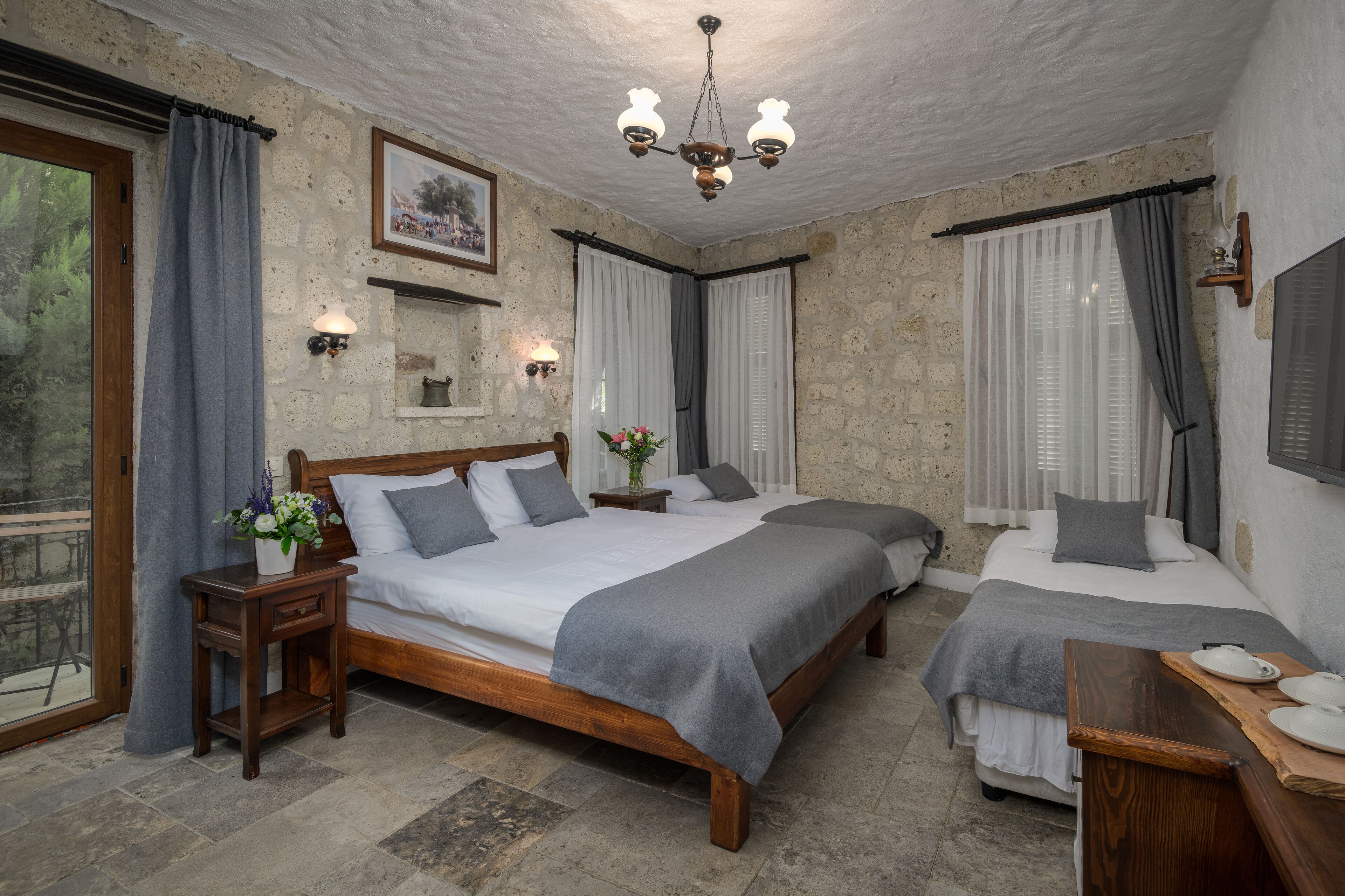 A room with two wooden beds, a chandelier, stone walls, and a view through a window.