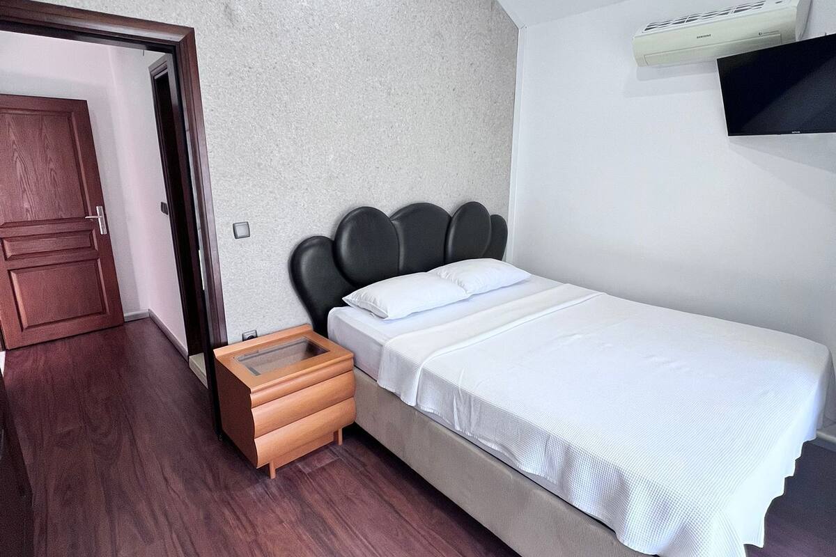 A bedroom with a bed, a wooden nightstand, a wall-mounted air conditioner, and a ceiling light fixture.