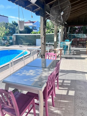 An outdoor pool area with a glass table and chairs, a hanging fan, and a covered seating area. - Dardanos Pansiyon (Çanakkale)