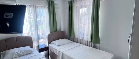 A hotel room with two beds, a flat-screen TV, a ceiling-mounted air conditioner, and a window with sheer curtains.