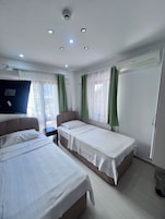 A hotel room with two beds, a flat-screen TV, a ceiling-mounted air conditioner, and a window with sheer curtains.