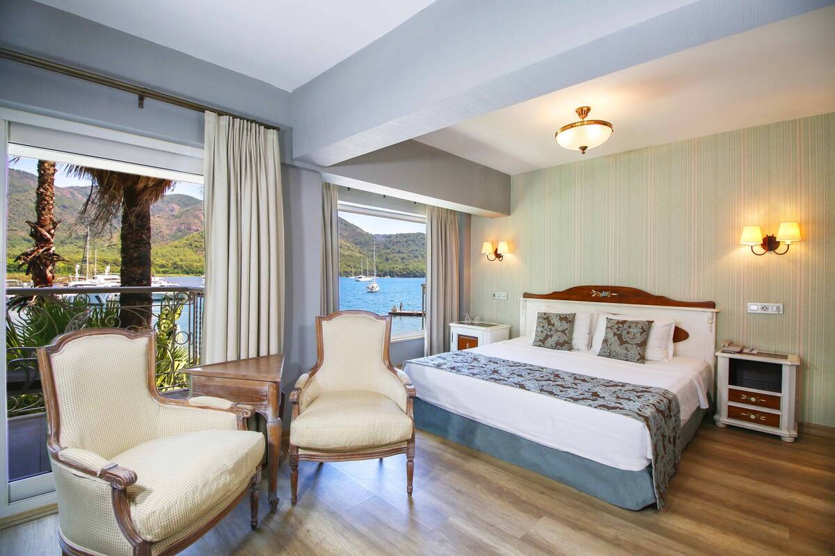 A hotel room with a bed, two chairs, a nightstand, a lamp, and a view of the sea.
