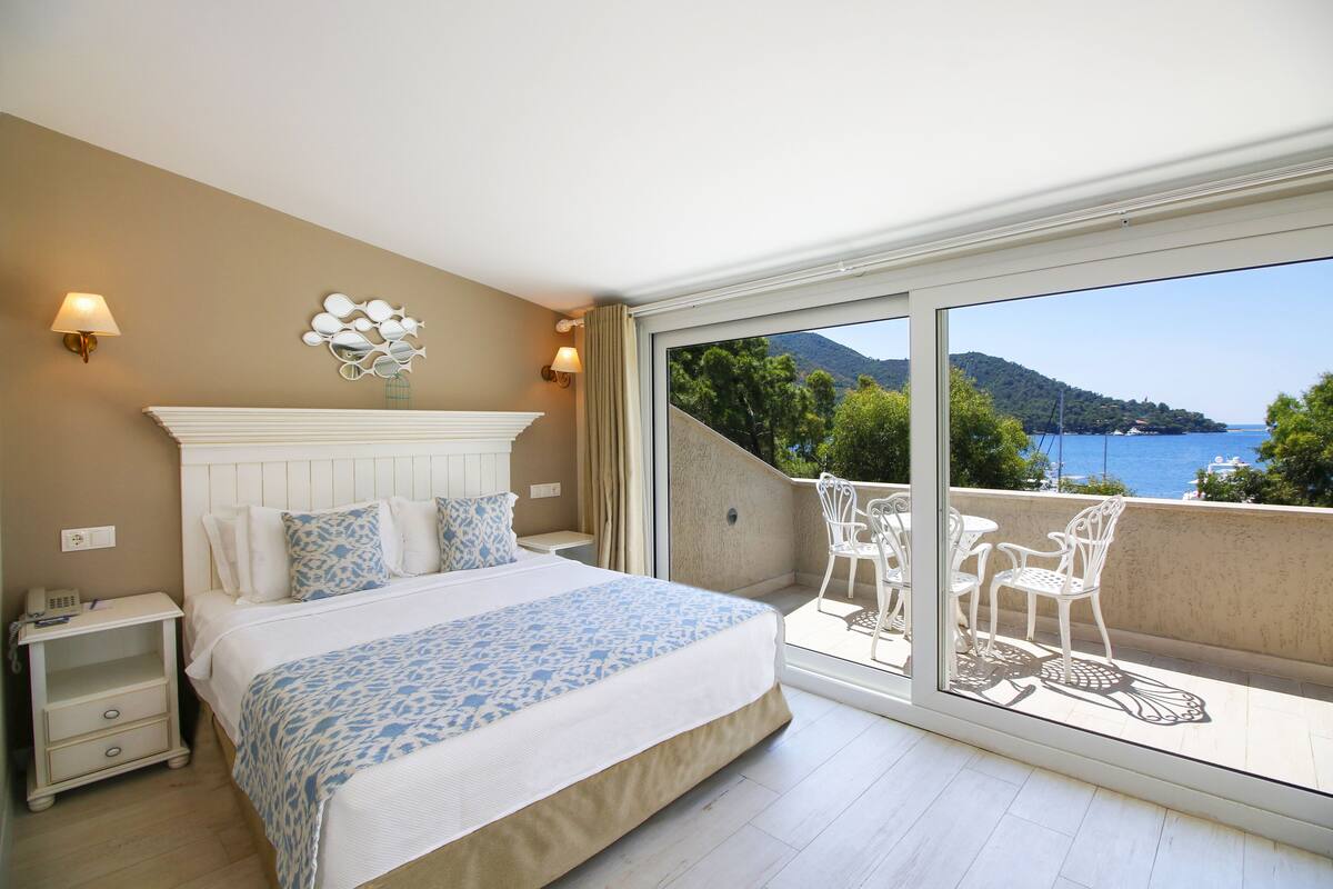 A hotel room with a large bed, a nightstand, a bedside table, a window with a view of the sea and mountains, and a balcony with outdoor furniture.