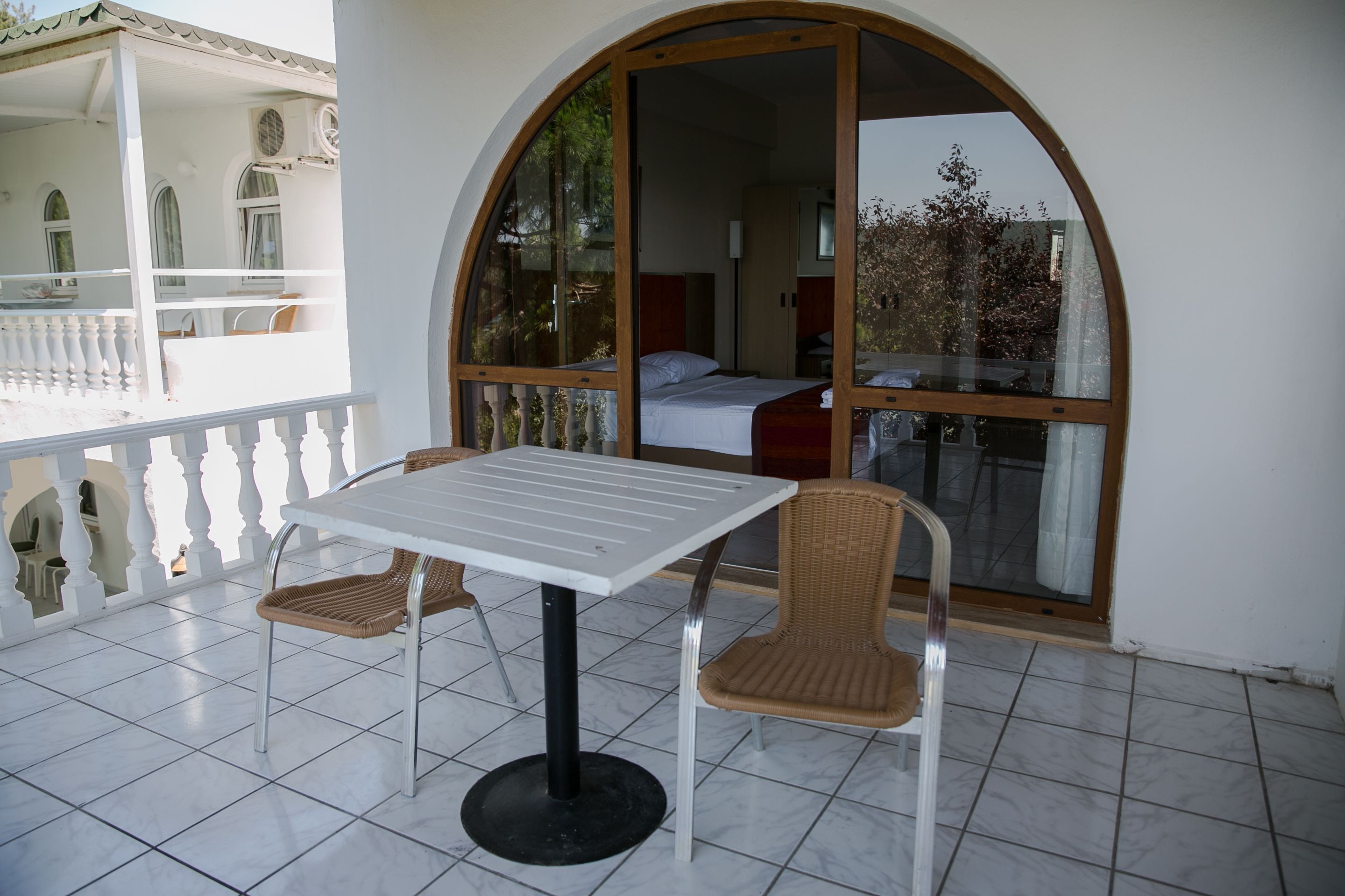 A balcony with a table and two wicker chairs, a large arched window, and a view of a bedroom with a bed and a nightstand.