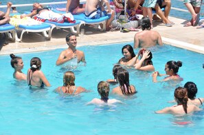 Una piscina con gente disfrutando del agua, sillas de descanso y una zona de terraza con baldosas.