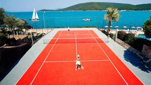 Una cancha de tenis con superficie roja, rodeada por una valla, con vista al mar y barcos al fondo.