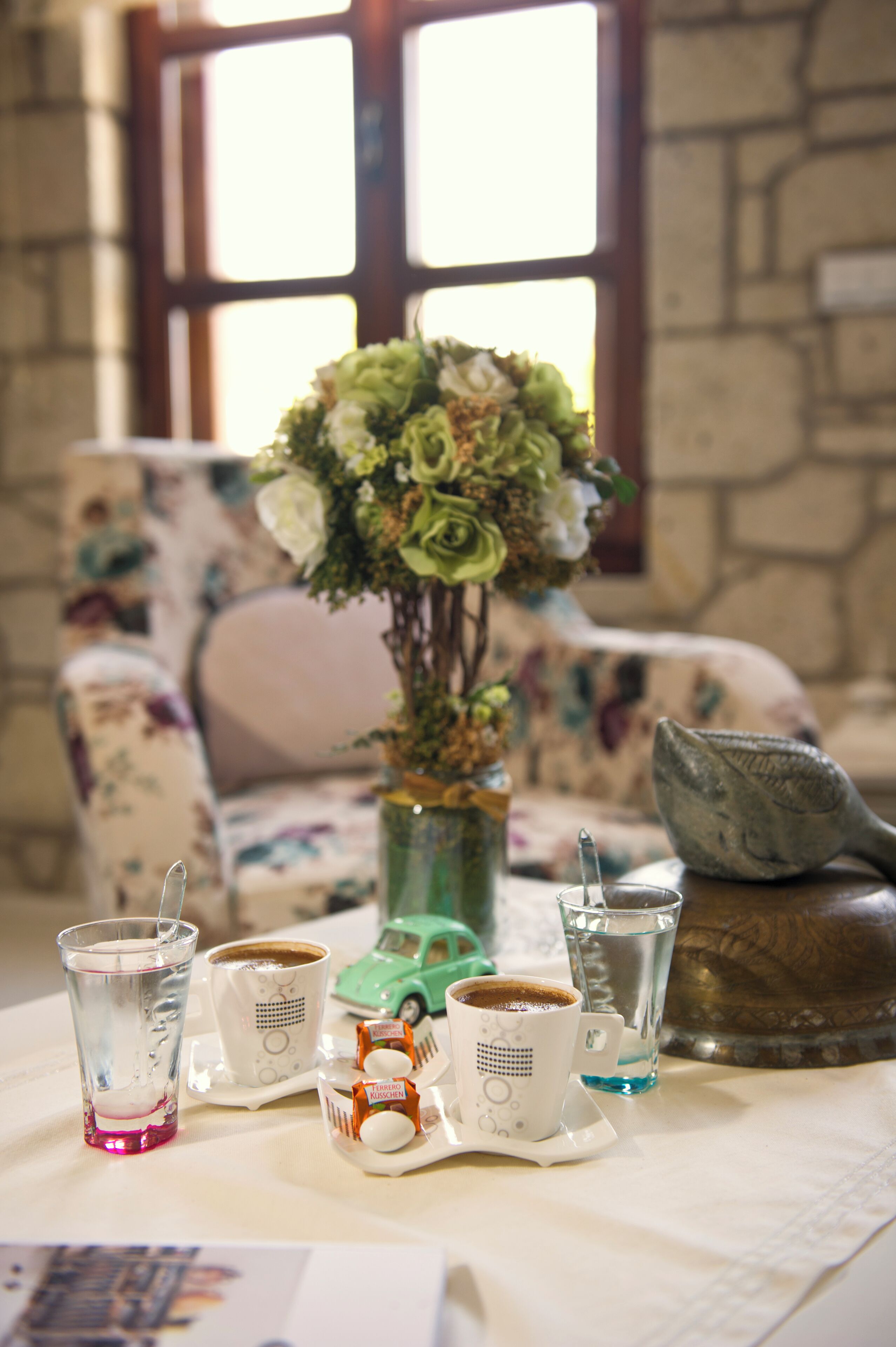 A table with a vase of flowers, two cups of coffee, a glass of water, and a small toy car.