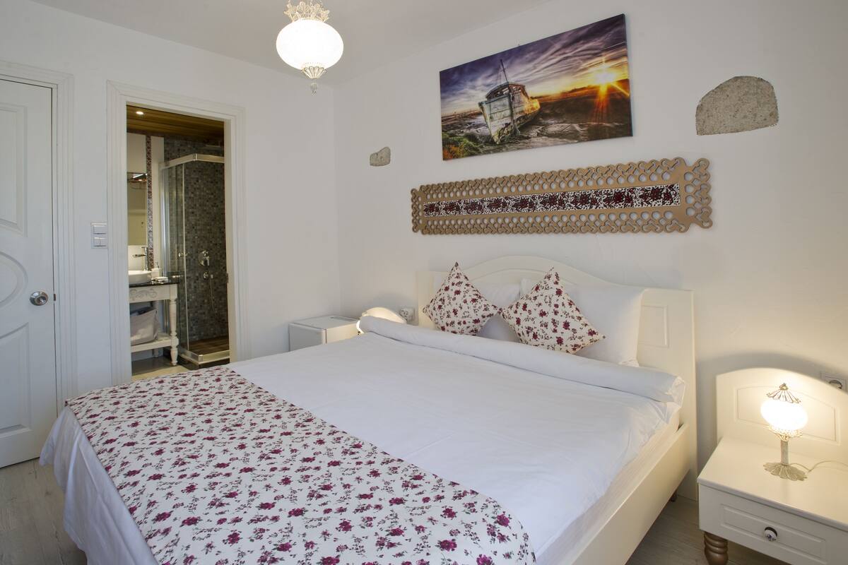 Un dormitorio ordenado con cama, mesitas de noche, una lámpara de pared y un cuadro de un atardecer.