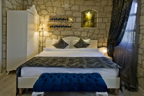 A bedroom with a stone wall, a bed with a patterned bedspread, a headboard with two decorative pillows, a bedside lamp, and a window with curtains. - Lodos Hotel (Cesme)
