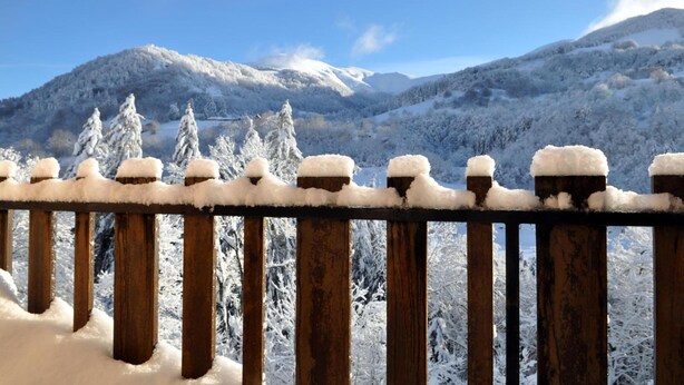 Una valla de madera con postes cubiertos de nieve y una vista de árboles y montañas nevadas al fondo.