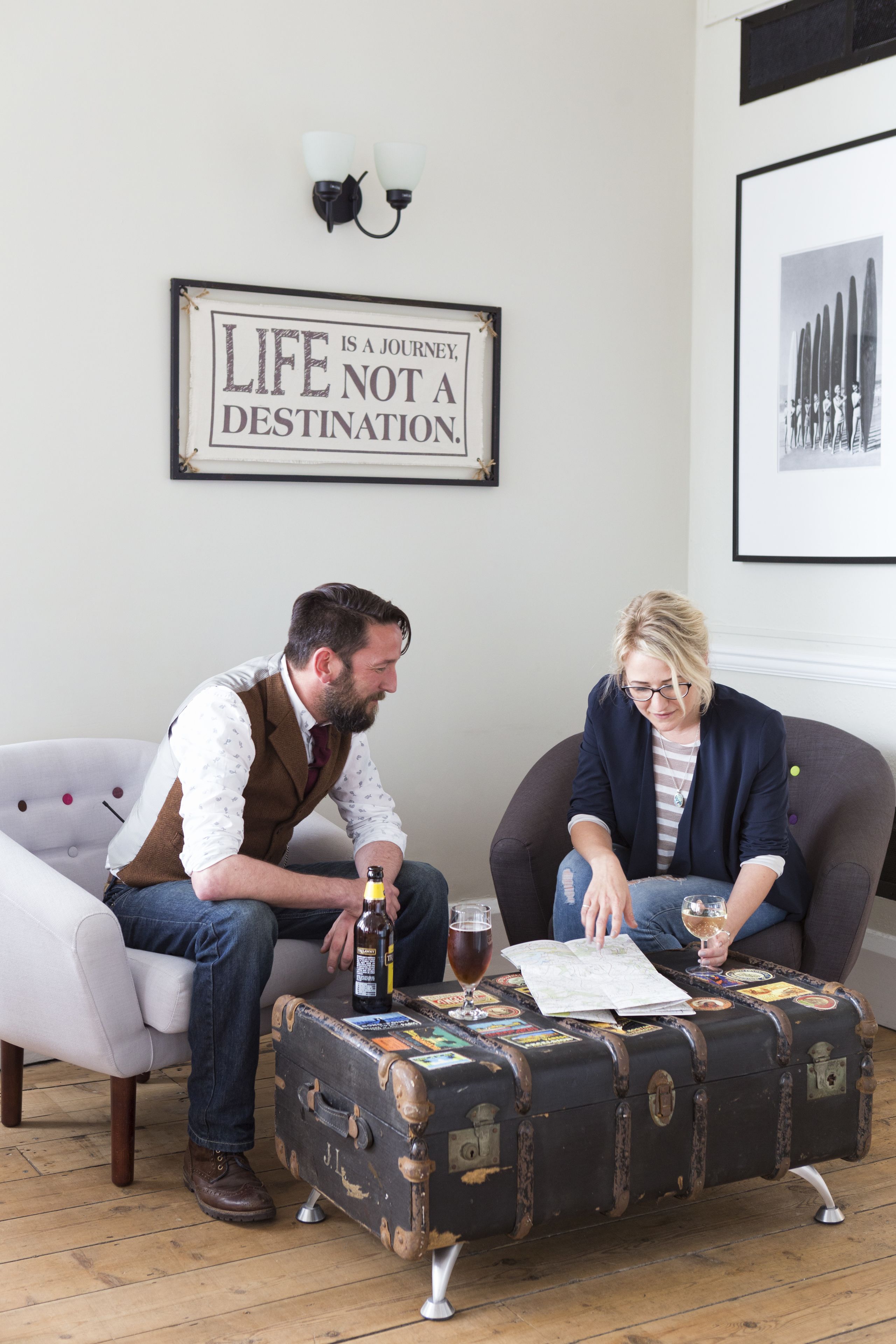 Zwei Personen sitzen auf einer Couch, dazwischen ein Beistelltisch mit einem alten Koffer, an der Wand ein gerahmter Spruch und darüber ein gerahmtes Bild.