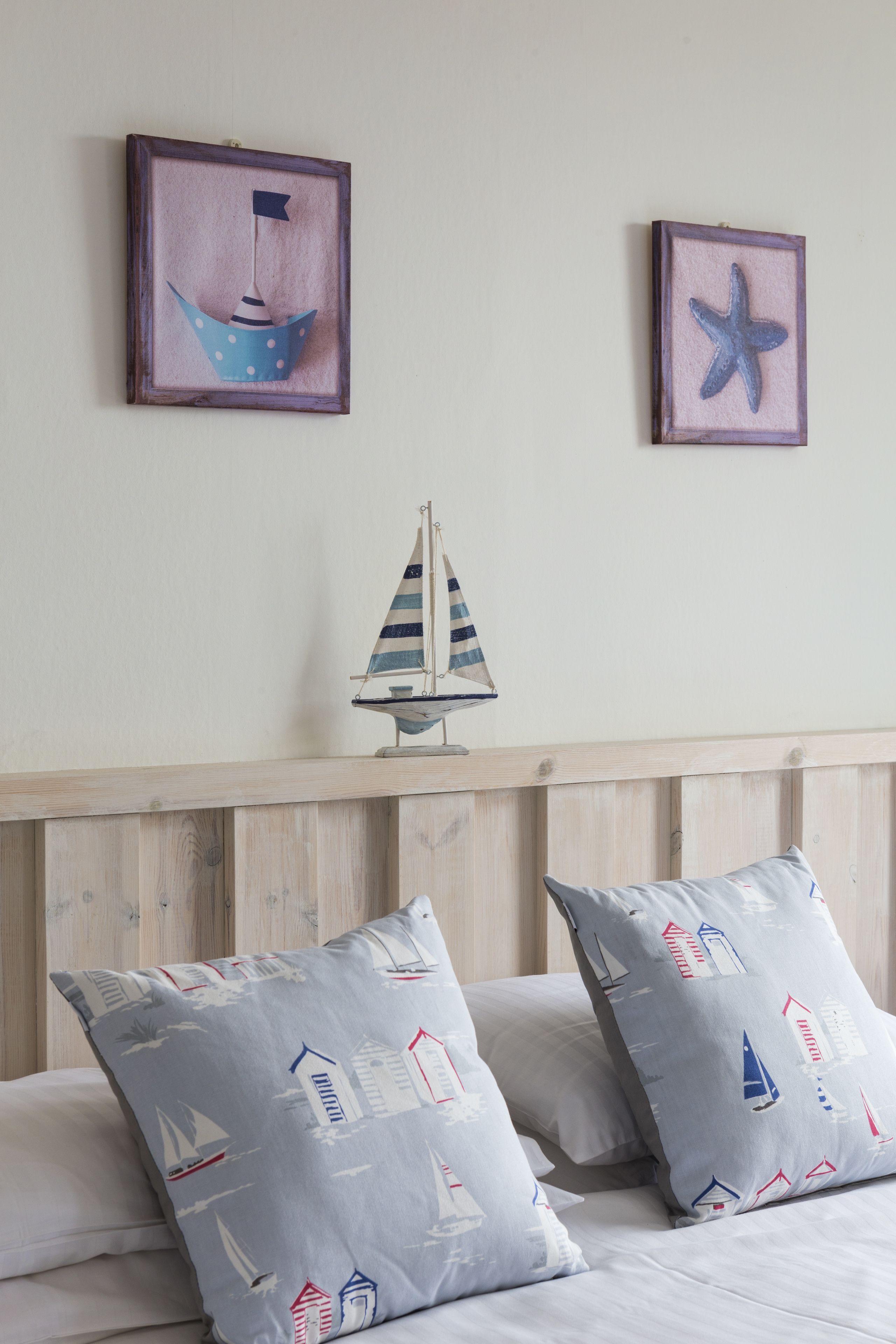 Un dormitorio con cabecera de madera, dos almohadas con motivos náuticos, una réplica de un pequeño velero y dos cuadros en la pared.
