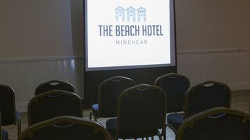 Una pantalla proyectando el logotipo de "THE BEACH HOTEL MINEHEAD" en un salón de conferencias con filas de sillas.