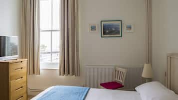 Habitación de hotel con una cama, un aparador de madera, una silla, una ventana con cortinas, un radiador y dos cuadros en la pared.