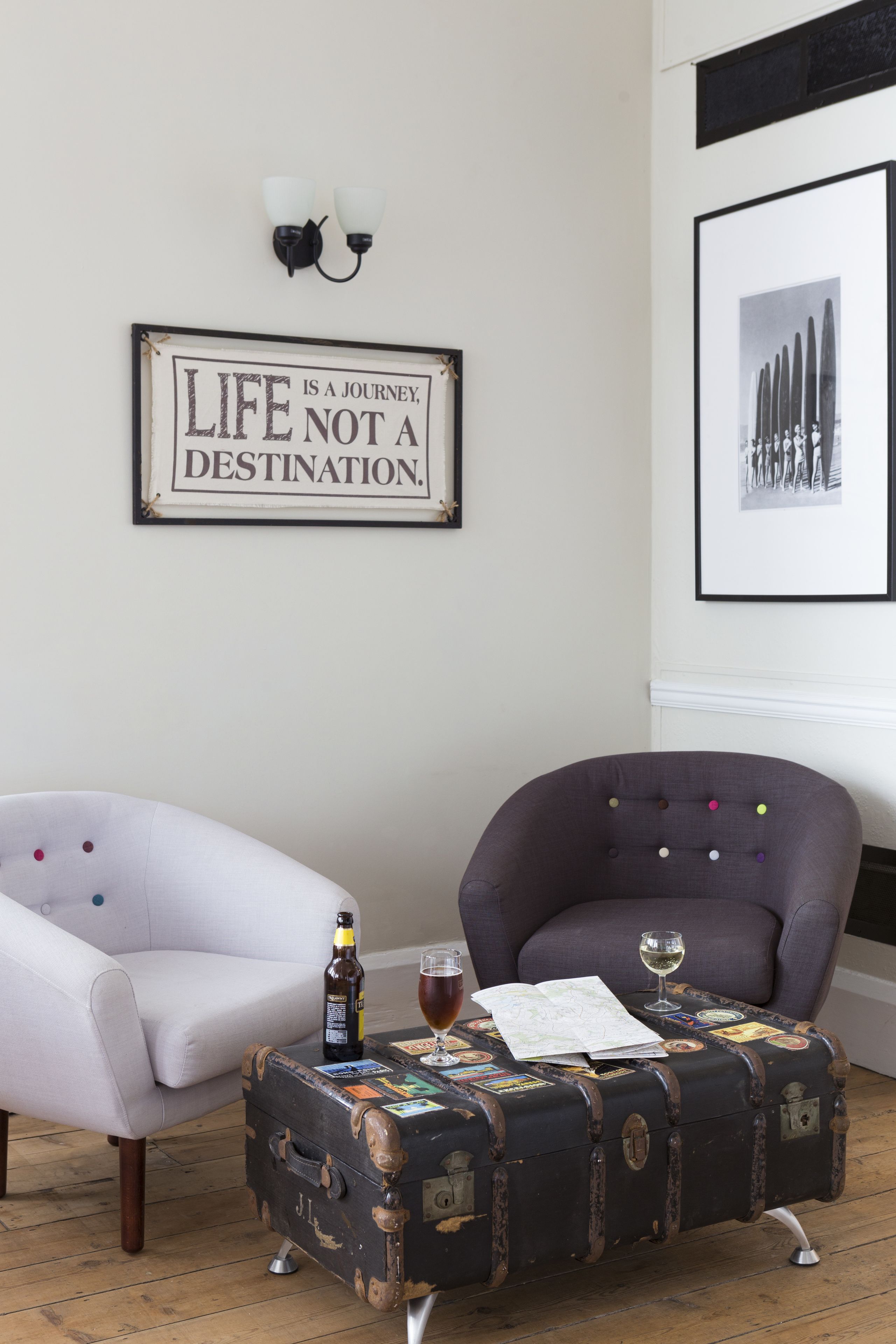 A room with a white armchair, a purple armchair, and a vintage suitcase table with a bottle and glass.