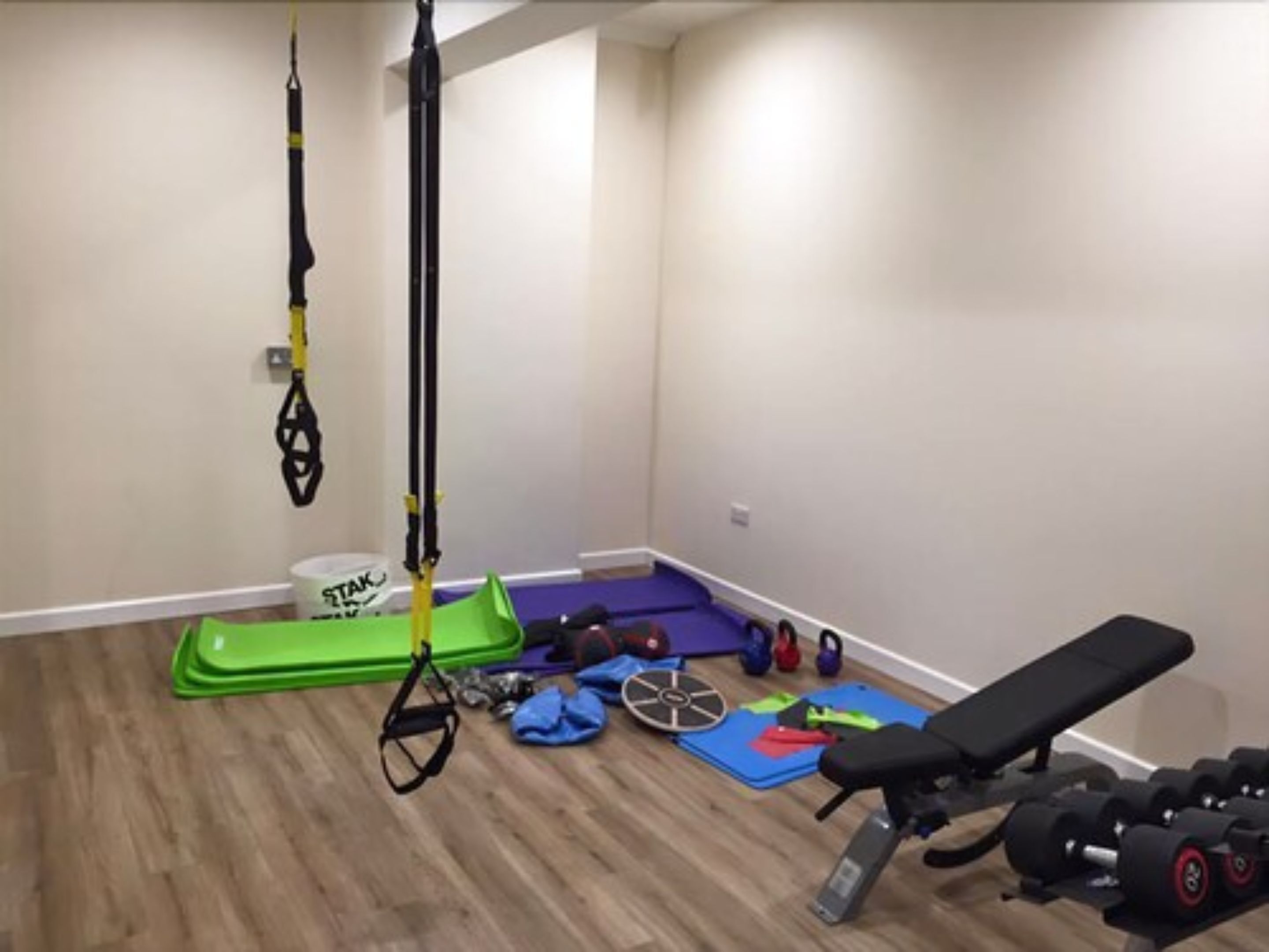 A small, sparsely furnished room with a wooden floor, a pull-up bar, a bench, and various exercise equipment.