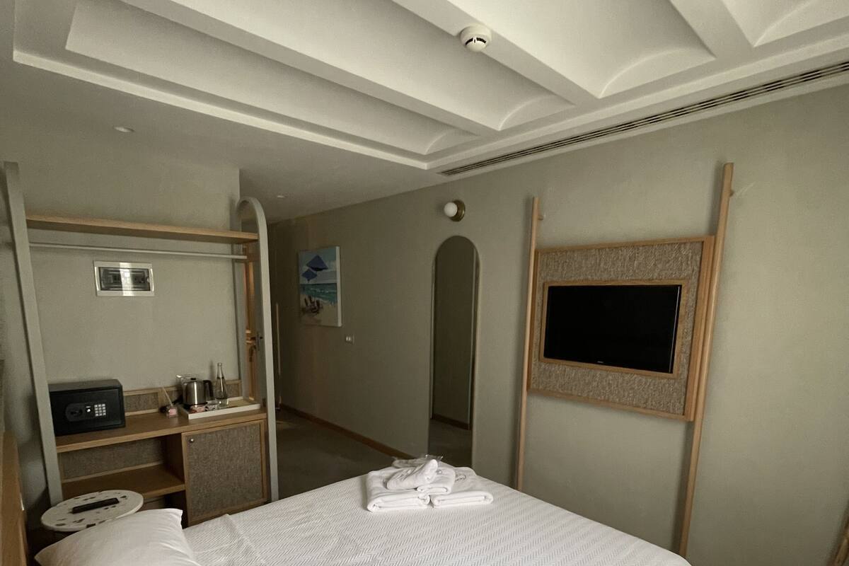 A hotel room with a bed, a desk with a computer, a mirror, a television, and a small kitchenette.