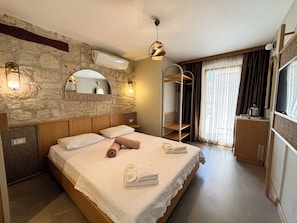 Hotelkamer met een stenen muur, een bed met witte lakens, een houten hoofdeinde, een hanglamp, een ingebouwde plank, een raam met gordijnen en een televisie op een standaard.