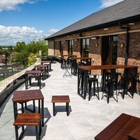 Un patio al aire libre con mesas y sillas de madera, un edificio de ladrillo con ventanas amplias, y una vista del cielo y las nubes.