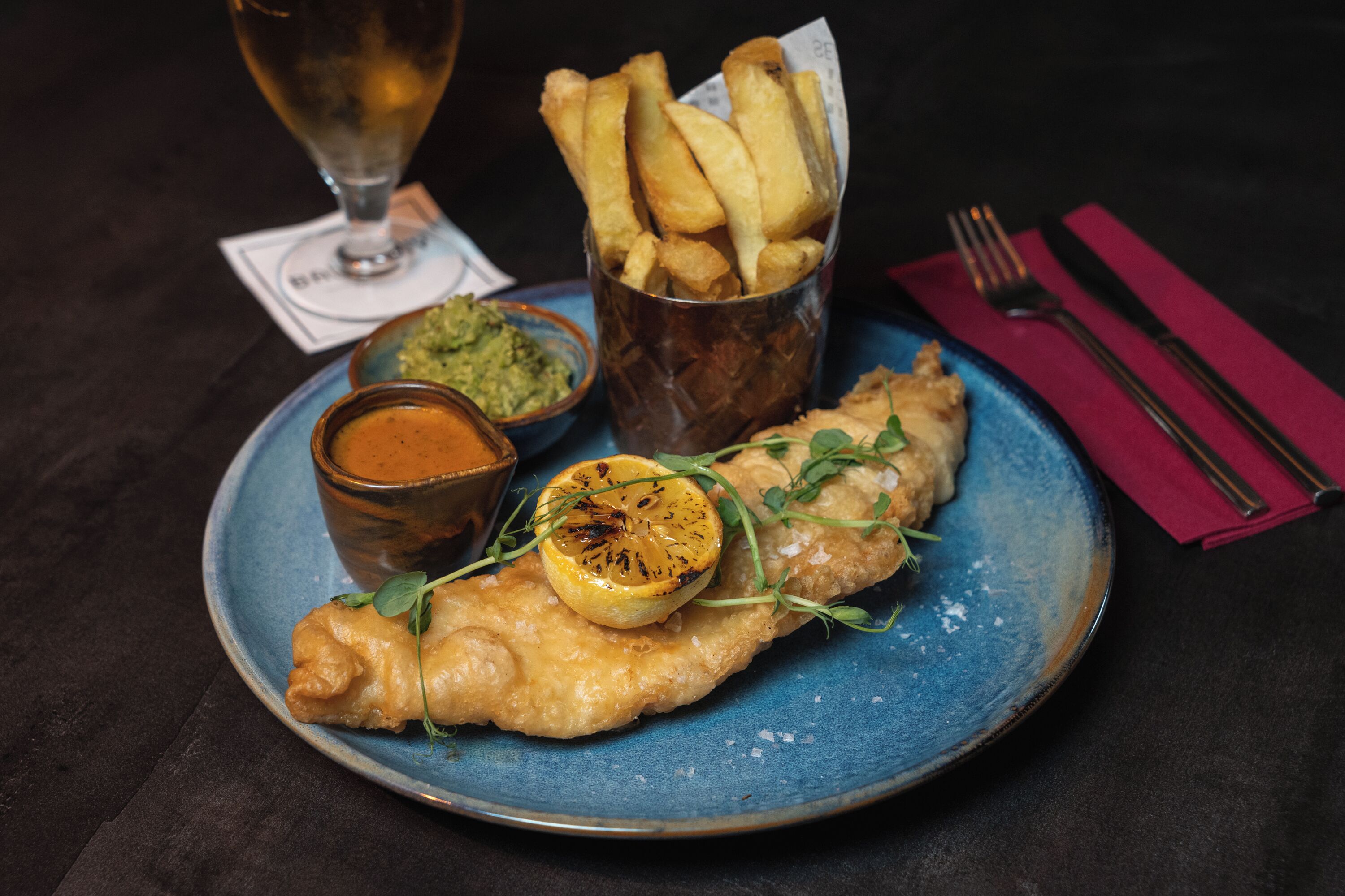 Une assiette de poisson-frites, un verre de bière et un petit bol de sauce.