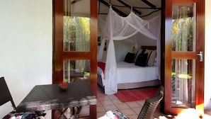 A room with a bed, a table with a chair, and a glass door leading outside. - Palmeiras Lodge (Vilanculos)