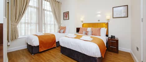 A hotel room with two beds, wooden flooring, and a large window with curtains.