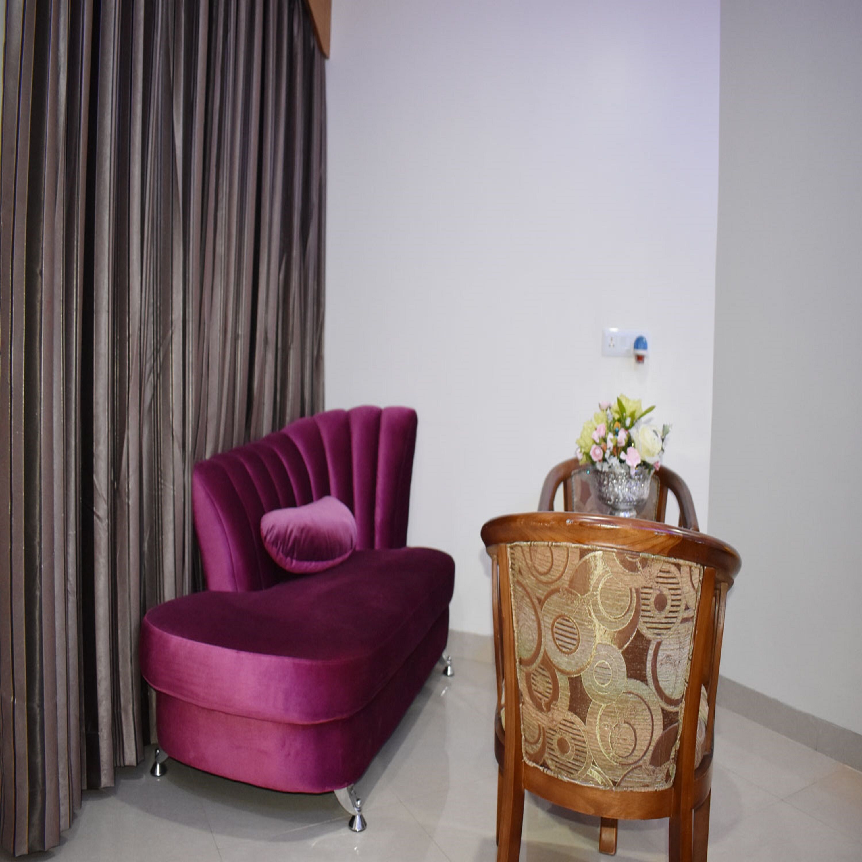 A room with a purple velvet sofa, a wooden chair with a patterned cushion, and a vase with flowers on a small table.