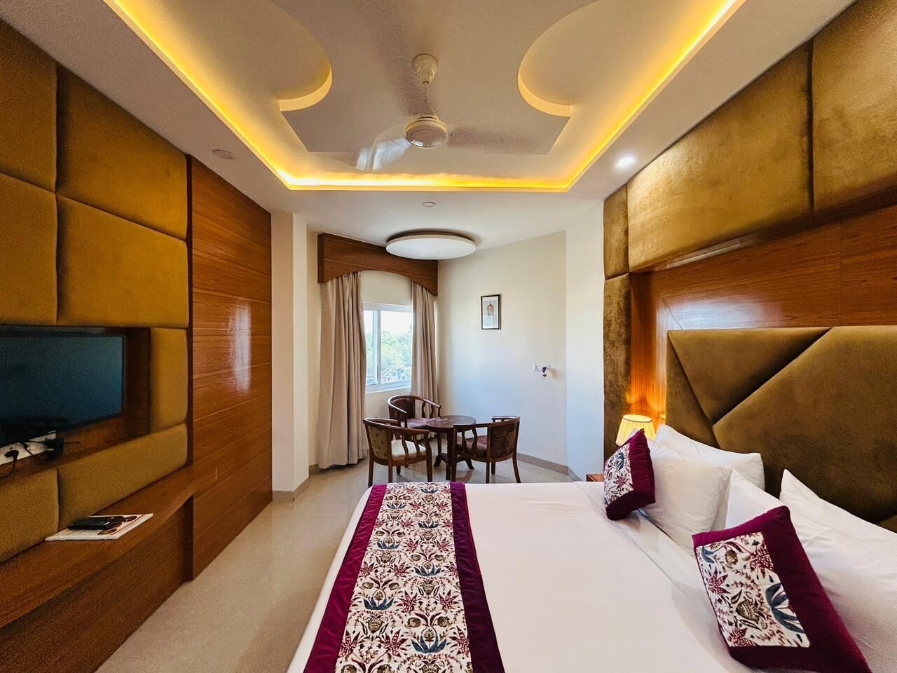 A hotel room with a bed, a television, a dining table with chairs, and a ceiling fan.