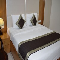 Una cama bien hecha con ropa blanca y dos almohadas decorativas, una lámpara de noche y una mesita de noche de madera con un cajón.